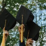 College graduates raise their caps together before a green tree.