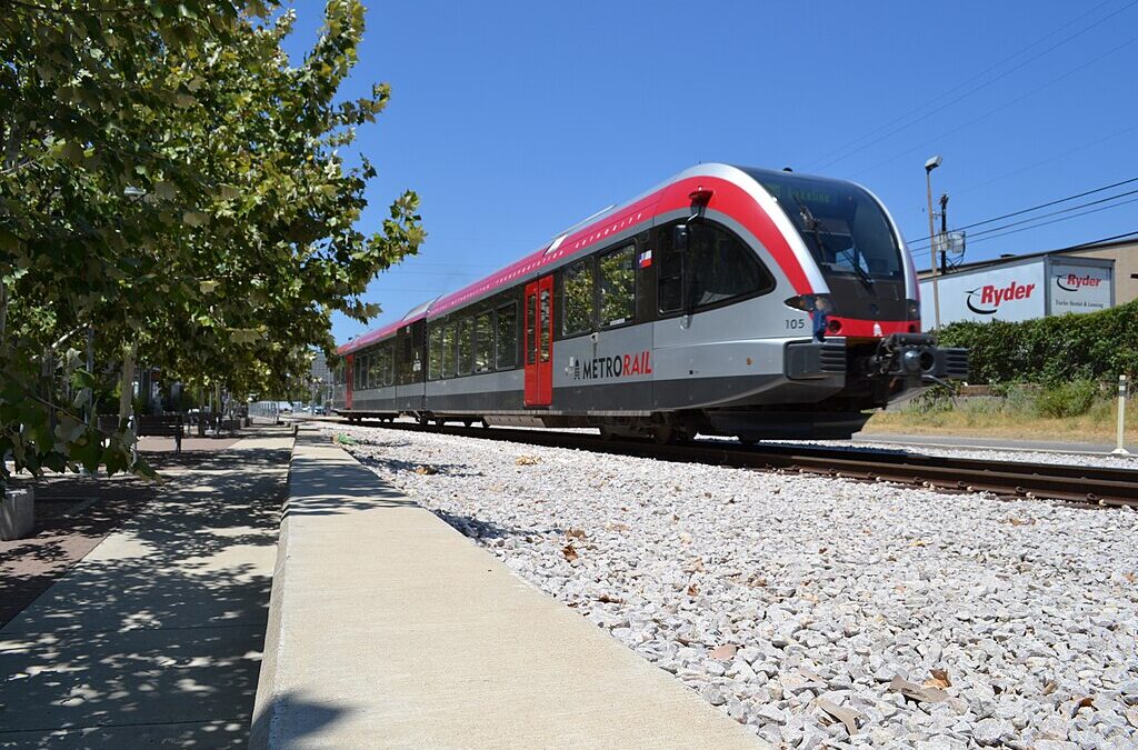 A CapMetro train along the tracks of a railroad near gravel and trees.