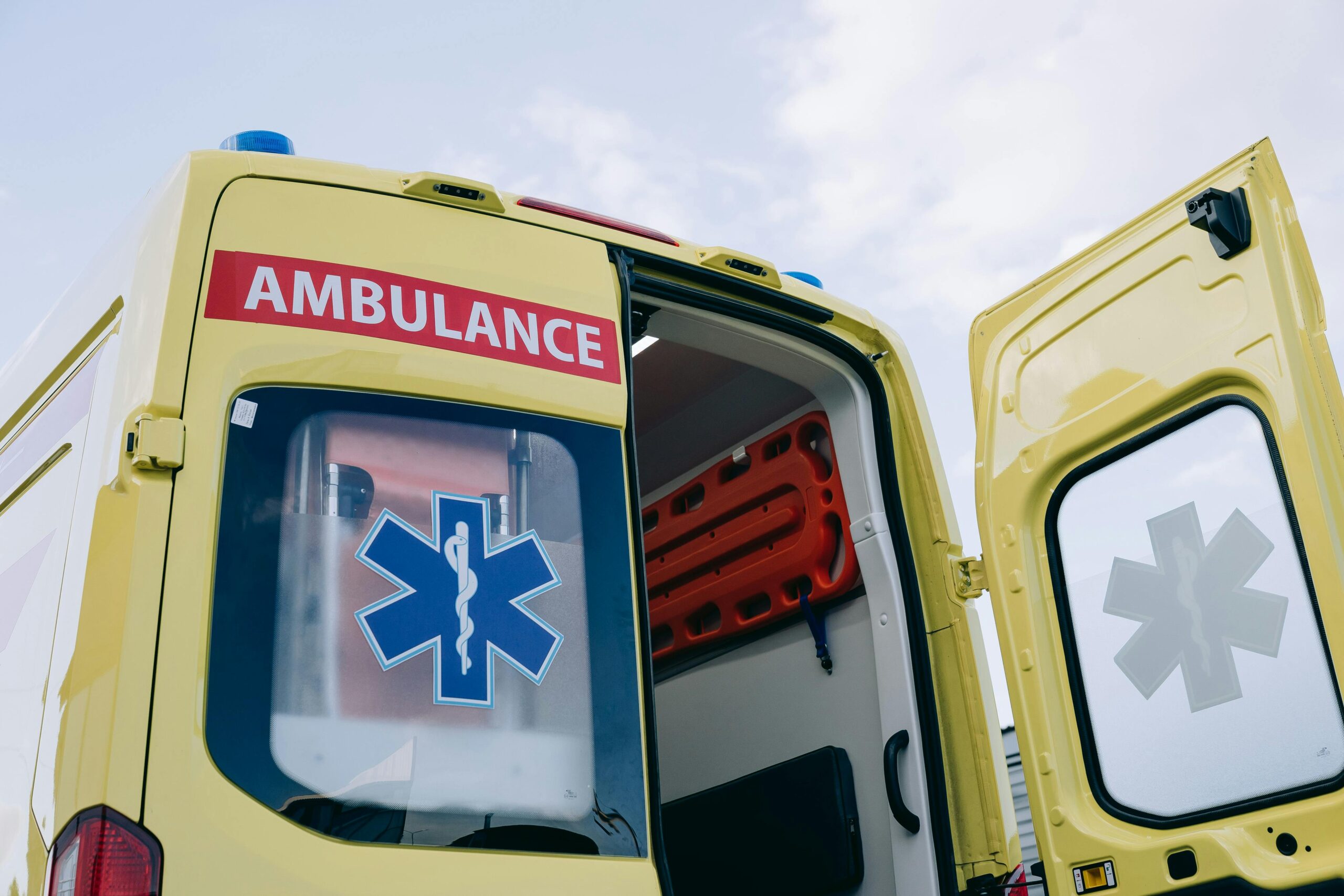 A yellow ambulance with one of the rear doors open before a cloudy sky.