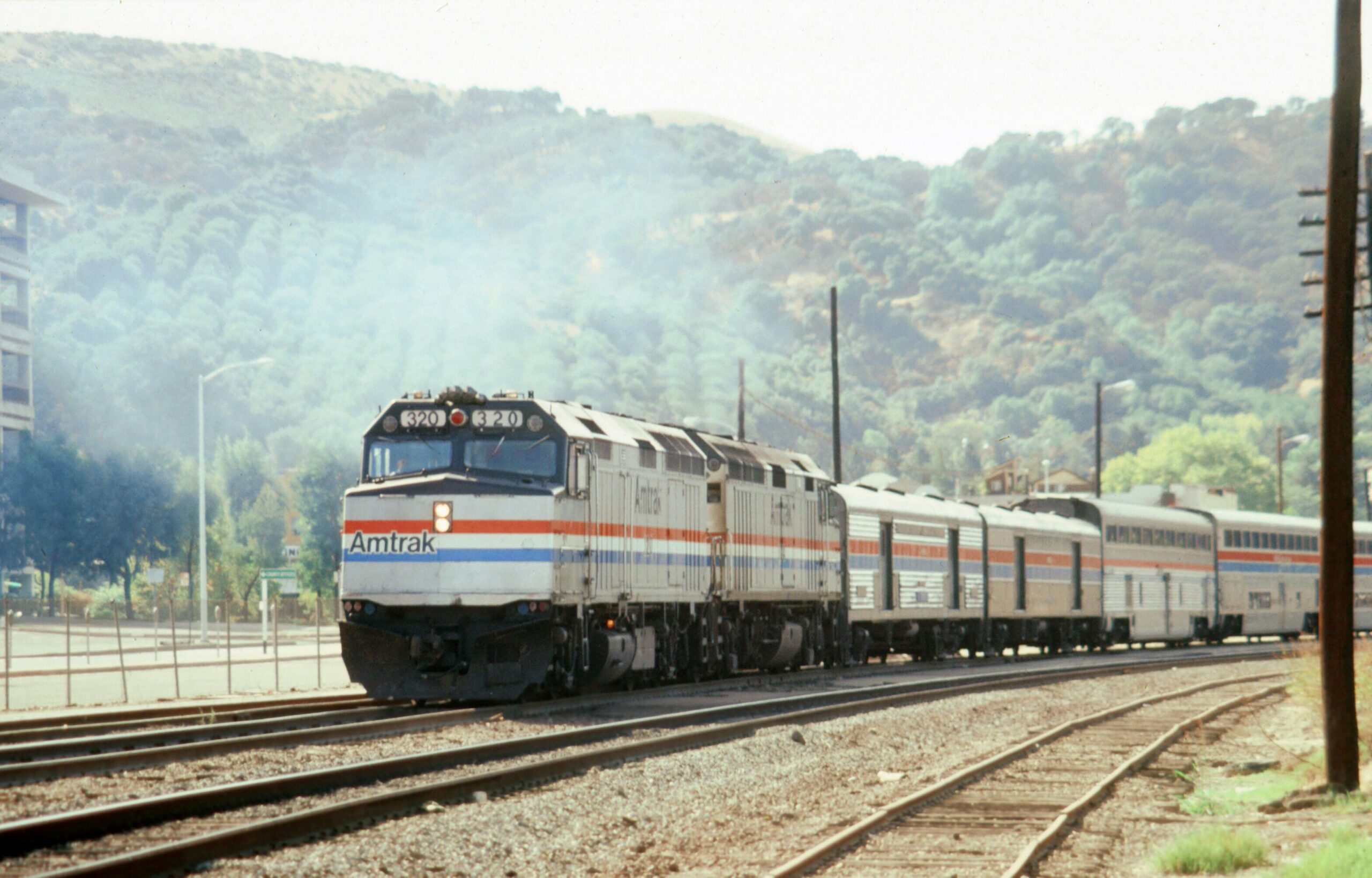 pexels-bill-silveira-2149789938-30892011 An Amtrak train on tracks before a green hill with trees and bushes.